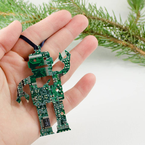 Hand holding a green robot-shaped keychain made from circuit board material against a white background with greenery.