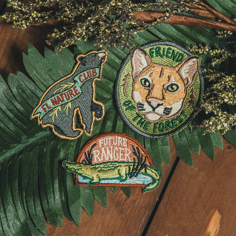 Three embroidered patches on a wooden surface with green leaves