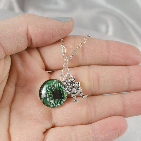 Hand holding a necklace with a green circuit board charm and a silver fairy pendant.