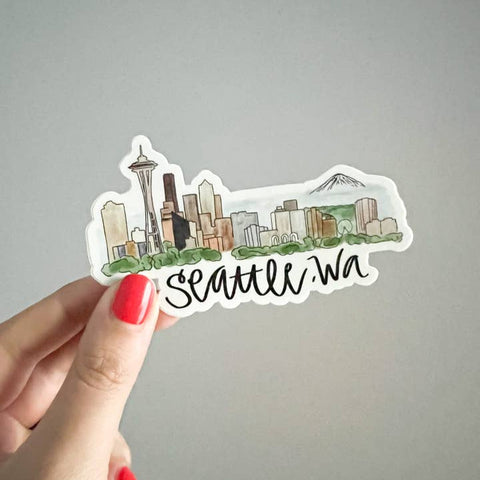 Sticker of Seattle, WA held by a hand with red nail polish on a gray background