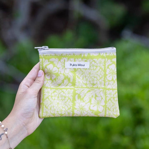 Hand holding a green patterned pouch with 'Puka Maui' label against a blurred green background