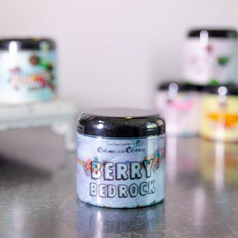 Jar of 'Berry Bedrock' candle on a reflective surface with blurred candles in the background.