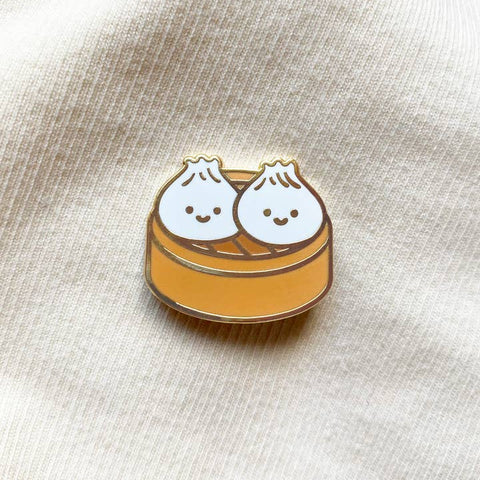 Pin with two cartoon dumplings in a steamer on a beige fabric background