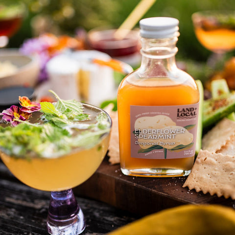 Bottle of Land & Local Elderflower Spearmint sparkling water on a table with cocktails and snacks.