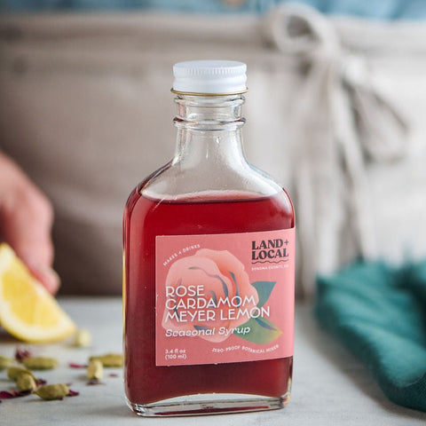 Bottle of Land & Local rose cardamom meyer lemon syrup on a table with a lemon and green leaves.