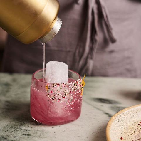 Gold bottle pouring liquid into a pink drink with a tea bag, on a marble surface.