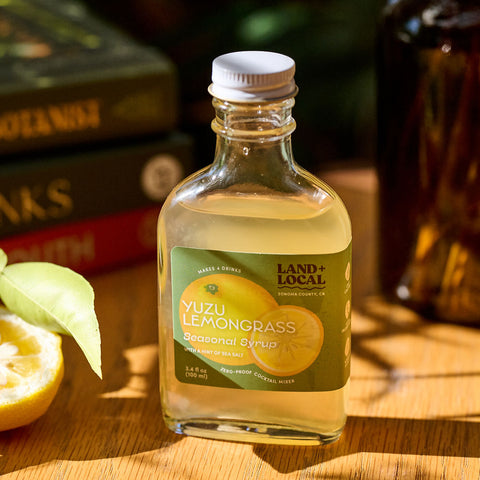 Bottle of Yuzu Lemongrass cocktail liquor on a wooden surface with lemons and books in the background.