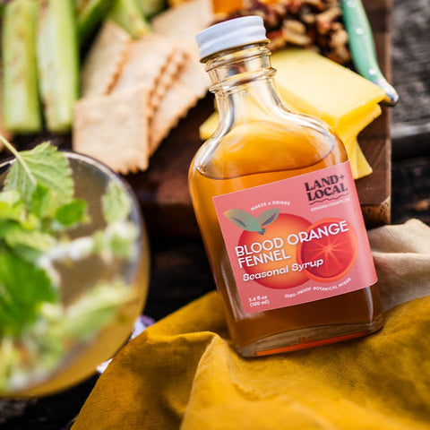 Bottle of 'Land Local' blood orange fennel syrup with a cocktail and garnishes on a table.