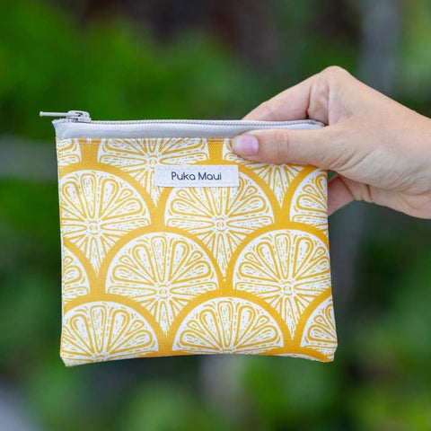 Hand holding a yellow patterned pouch with 'Puka Maui' label against a blurred green background
