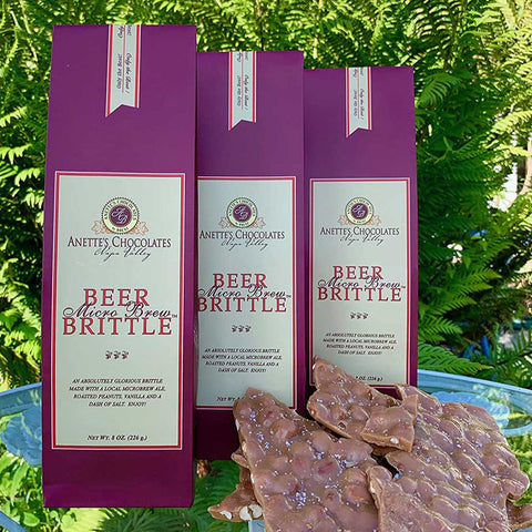 Three packages of Anette's Chocolates Beer Alewe Brew Brittle with brittle pieces on a green leafy background.