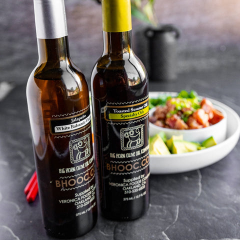 Two bottles of aged balsamic vinegar, one labeled as 'White Balsamic' and the other as 'Tuscan Balsamic', with a dish containing food items in the background.