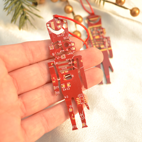 Red circuit board design hanging ornament held in a hand with Christmas tree decorations in the background.