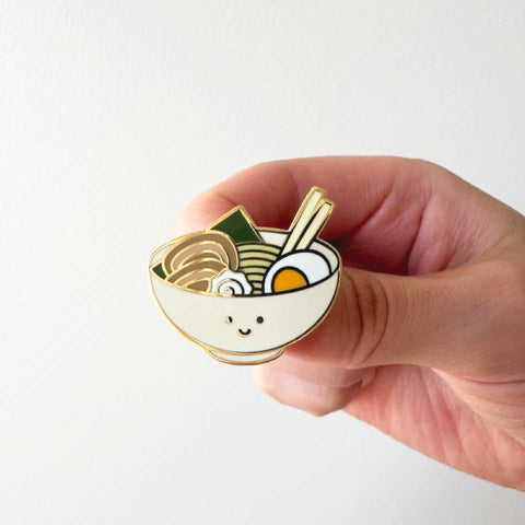 Hand holding a cute enamel pin of a bowl with noodles, eggs, and vegetables on a white background