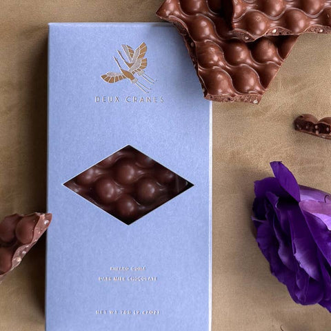 Blue chocolate box with a gold logo on a brown surface with chocolate pieces and purple flowers.