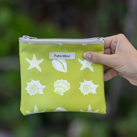 Hand holding a green pouch with white shell patterns against a blurred natural background