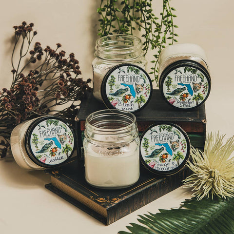 Candles in glass jars with decorative labels on a wooden stand, surrounded by plants.