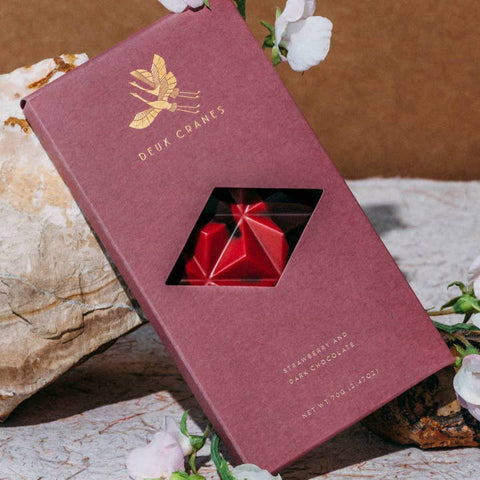 Red jewelry box with a gemstone inside, branded 'Deux Carres', on a natural stone surface with flowers.