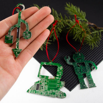 Hand holding a green circuit board ornament with more ornaments on a black surface.
