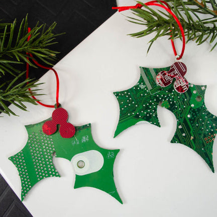 Green holly ornament made from recycled computer circuit board with red berries on a white surface with Christmas tree branches.