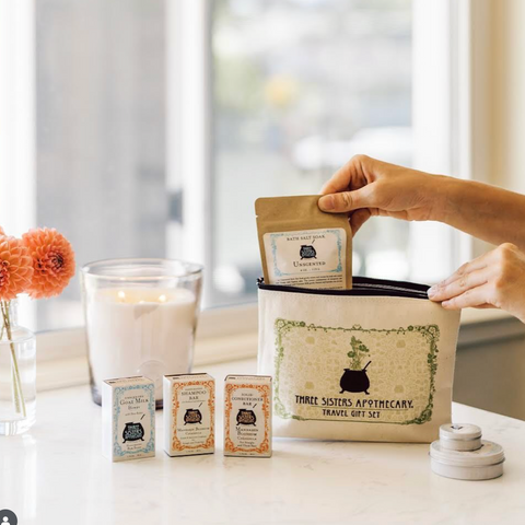 Three Sisters Apothecary travel gift set with a hand holding a package on a table.