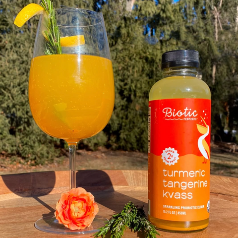 Glass of orange drink with garnish next to a bottle of Biotic Turmeric Tangerine Kvass on a wooden surface outdoors.