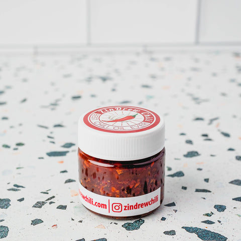 A small, travel-sized container of Zindrew Crunchy Garlic Chili Oil placed on a surface with a white tiled background and a red and white label.