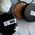 A close-up of a skincare product labeled 'SWASH THERAPY SOUDOR' on a black tin, with one tin open to show a beige cream and another closed lid nearby on a light surface.