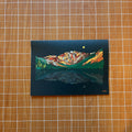 Artistic depiction of a mountain landscape on a dark background, placed on a wooden surface with grid lines.