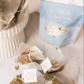 Tea bags and a box of Leaves of Leisure 'Snow Angel' herbal tea arranged on a light marble surface next to a white ribbed ceramic container.