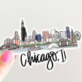 Sticker of Chicago skyline with 'Chicago, IL' text held by a hand on a light gray background