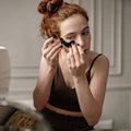 A woman with red hair in a bun applies a black eye patch while looking into a mirror. She is wearing a dark brown sports bra and shorts in a brightly lit room with white walls.