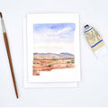 A watercolor landscape art print on a white card, styled in a flat-lay with a paintbrush on the left and a tube of paint on the right, on a clean white background.