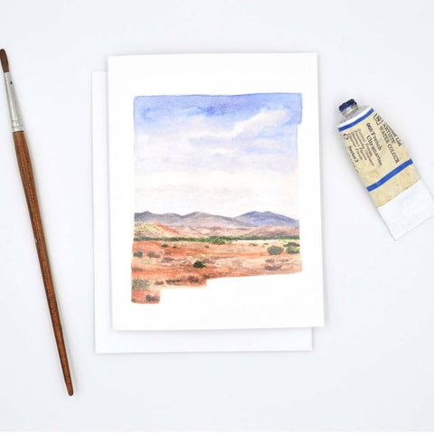 A watercolor landscape art print on a white card, styled in a flat-lay with a paintbrush on the left and a tube of paint on the right, on a clean white background.