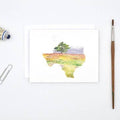 A watercolor art print of Texas showing a tree in a field, displayed on a white background with a paint tube, paperclip, and paintbrush nearby.