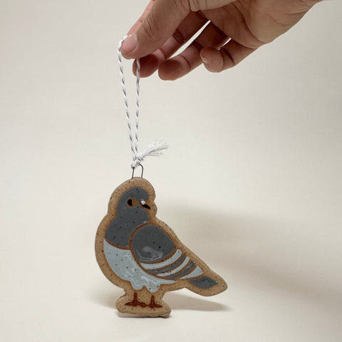 A hand holds a small bird-shaped ornament or charm with a white string hanger against a plain light background. The bird is painted gray, white, and brown with a tan outline, and the hand is shown from above.