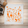 White towel with orange pelican designs on a wooden surface with a cup of coffee.