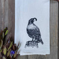 White towel with black bird design on a wooden surface with plants.