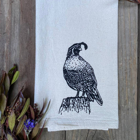 White towel with black bird design on a wooden surface with plants.