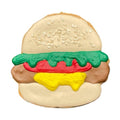 A sugar cookie shaped like a hamburger, decorated with light brown icing for the bun, green, red, yellow, and brown frosting layers, and white icing sesame seeds on the top bun, isolated on a white background.