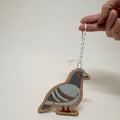 A hand holds a small wooden pigeon ornament with a white hanging string against a plain light background. The ornament is made from natural wood with painted gray, blue-gray, and white details, plus small brown and red accents.