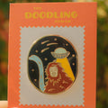 Enamel pin with a design of a person with a helmet and a crescent moon, held on an orange card with 'The Doodling Nomad' branding.