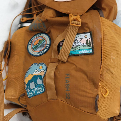A tan waterproof backpack with multiple embroidered outdoor patches, shown resting on white snow. The backpack features a drawstring top, compression straps, and various patches including 'Alpine 267', 'Washington', and 'Biking'.