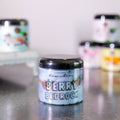 Jar of 'Berry Bedrock' candle on a reflective surface with blurred candles in the background.