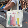A person carrying a large cream canvas tote bag featuring a colorful 'NEW YORK CITY' skyline print with skyscrapers, taxis, and the Statue of Liberty, photographed on a city street.