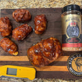 Crispy glazed meat wings arranged on a wooden cutting board with two seasoning bottles, a yellow thermometer, and King Louie's branding on a granite countertop.