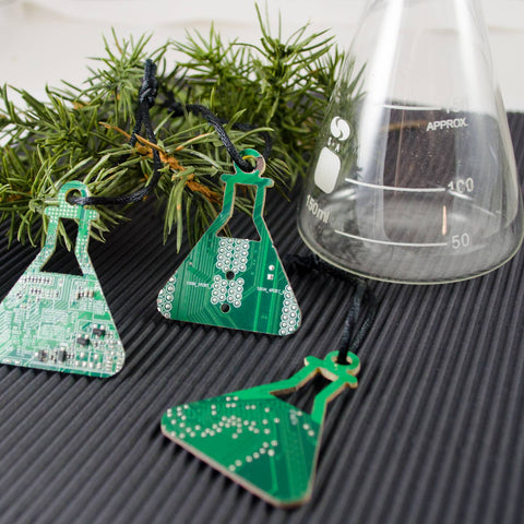 Green beaker-shaped earrings on a textured surface with a branch and beaker in the background.