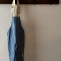 A blue denim bib apron with cream-colored shoulder straps hangs from a dark wooden wall hook on a plain beige wall.
