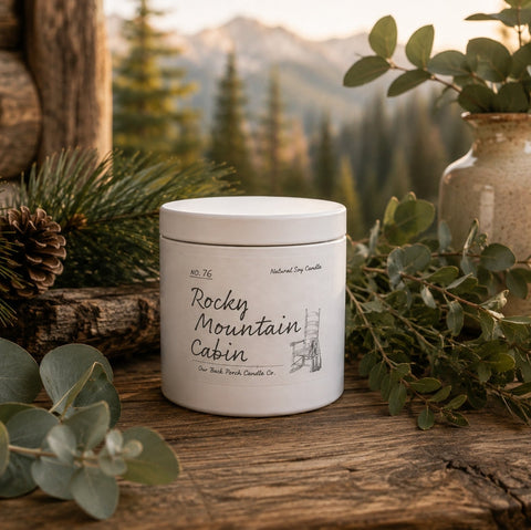 A white jar candle labeled 'Rocky Mountain Cabin' sits on a rustic wooden surface outdoors, surrounded by eucalyptus leaves, pine branches, a ceramic vase with greenery, and a lantern, with a mountain forest background.
