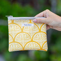 Hand holding a yellow patterned pouch with 'Puka Maui' label against a blurred green background