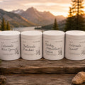 Four white metal tins of natural body care products lined up on a rustic wooden surface with rocks and evergreen branches, set against a scenic mountain lake at sunset.